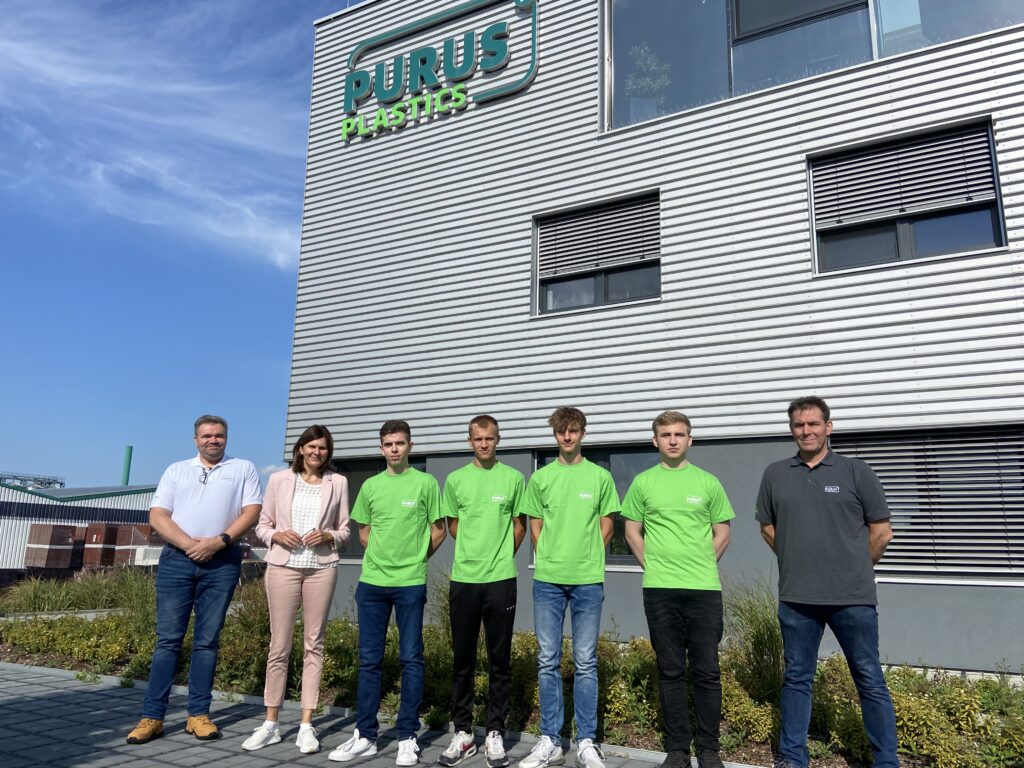 Sieben Personen in grünen, grauen und weißen Shirts stehen nebeneinander vor dem PURUS PLASTICS Verwaltungsgebäude und schauen in die Kamera