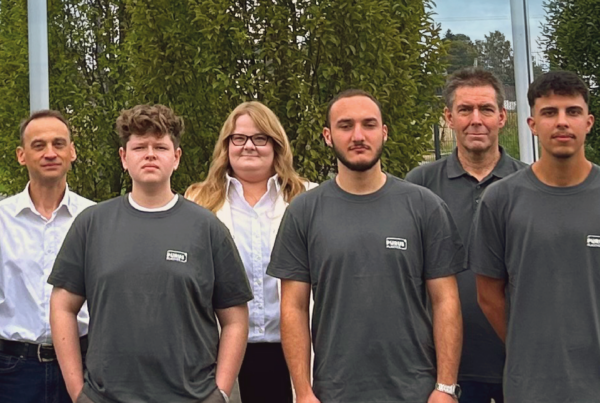 Acht Personen in grauen Shirts und weißen Hemden stehen nebeneinander auf einem Parkplatz und schauen in die Kamera