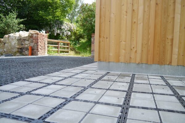 Kombination aus ECORASTER Bloxx und ECORASTER mit Schotter verfüllt als Außenfläche einer Scheune mit Natur und Grün im Hintergrund