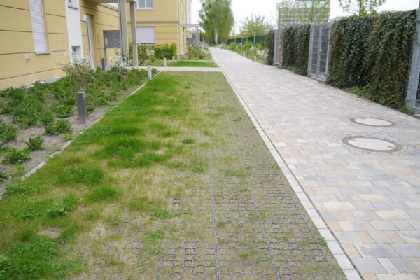 Begrünte ECORASTER Feuerwehrzufahrt entlang eines gelben Wohnkomplexes und gepflastertem Weg