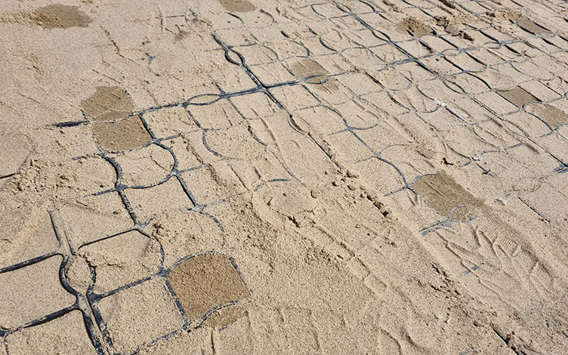 Ansicht von oben auf Sand mit Wasserflecken. Das Ecoraster E40 Aqua schaut an manchen Stellen hervor.